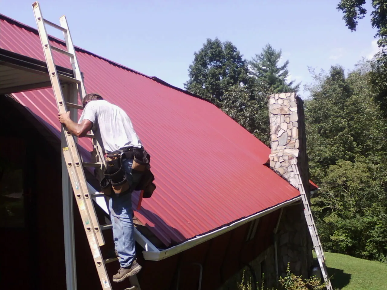 Roofer on a ladder beside a red metal roof and stone chimney — Soffit & Fascia Repair by Roofing Brothers in Union Point